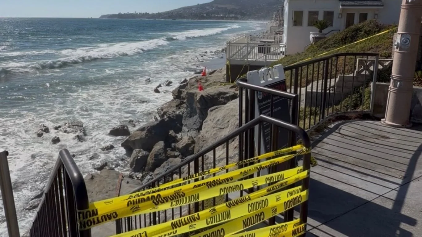 Caution tape across a closed beach stairway above surf and exposed rock at Brooks Street in Laguna Beach, California