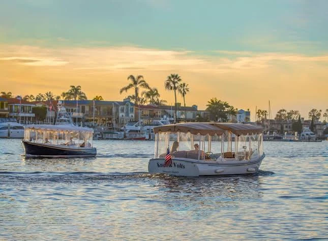 Two Duffy electric boats cruising past bayfront homes on Newport Harbor at golden hour in Newport Beach, California